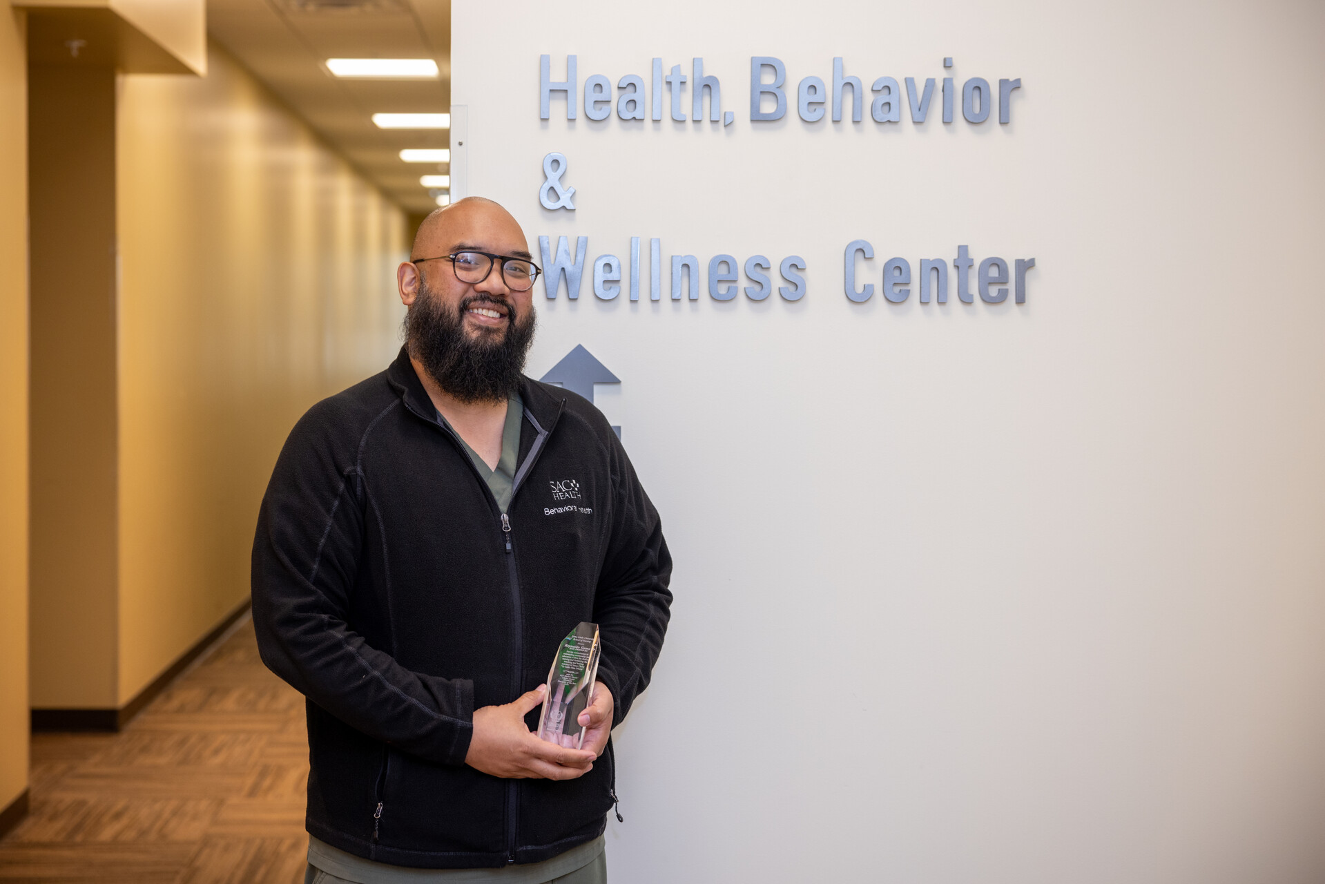 A bearded healthcare employee wearing glasses stands in a hallway holding a clear award plaque beside a wall sign reading “Health, Behavior & Wellness Center.” The person smiles at the camera while wearing dark medical attire and a black zip-up jacket. The hallway extends into the background with soft overhead lighting.