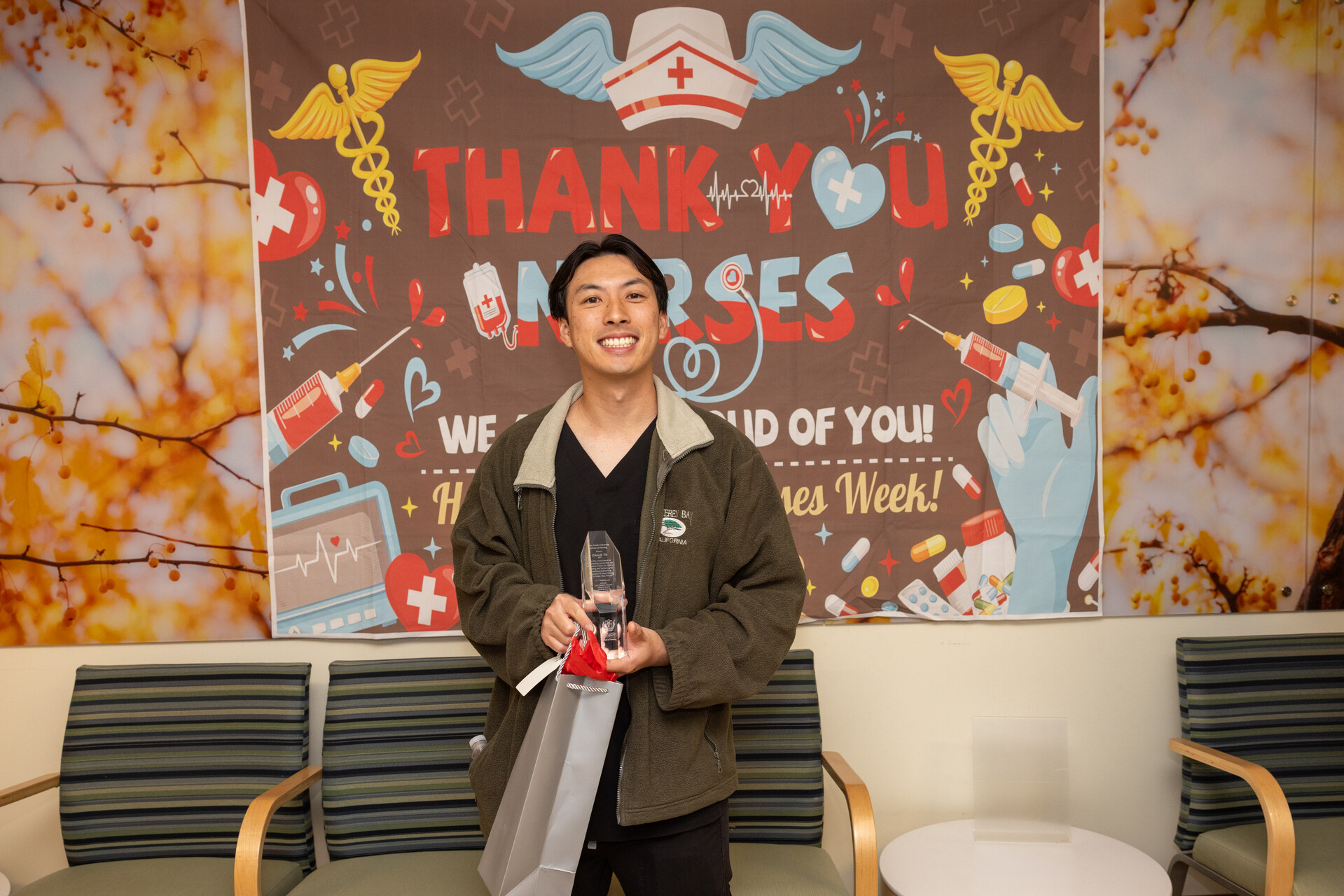 A smiling healthcare worker stands indoors holding a glass award and a gift bag in front of a large colorful “Thank You Nurses” banner decorated with medical symbols, syringes, pills, and hearts. The person wears dark scrubs and a zip-up jacket and faces the camera in a waiting-room style space with chairs on both sides.