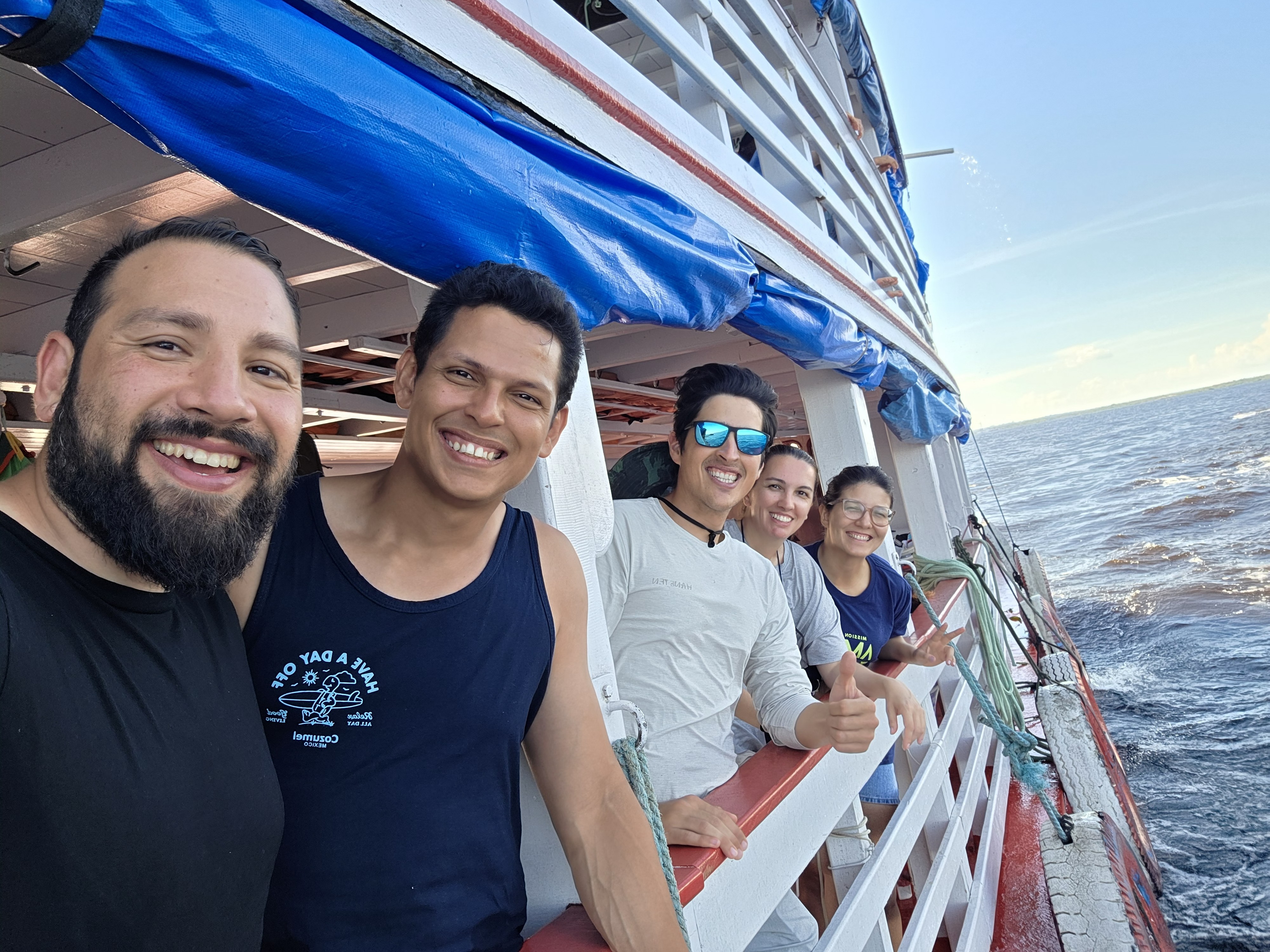 A group of five mission trip volunteers consisting of students, alumni and administration smile and stand on the side of a boat traveling the Amazong River in Brazil.