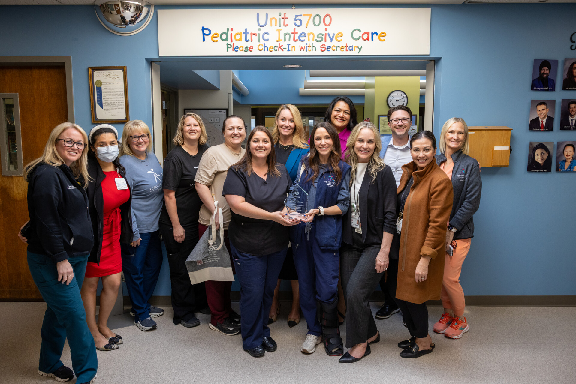 A group of LLU Nursing professionals and leaders stand beneath a sign reading “Unit 5700 Pediatric Intensive Care” posing for a group photo in a hospital corridor, celebrating the unit receiving a Nurses Week honor.
