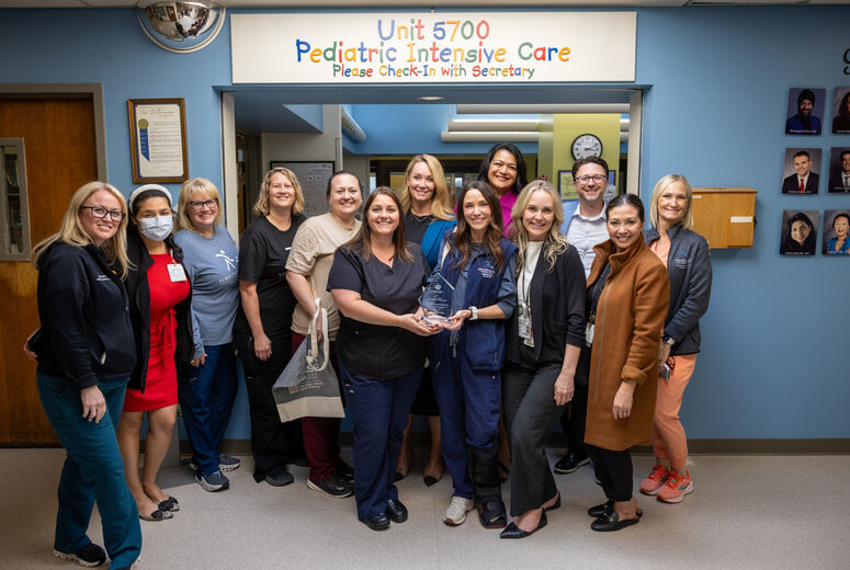 A group of people stand beneath a sign reading “Unit 5700 Pediatric Intensive Care” posing for a group photo in a hospital corridor.