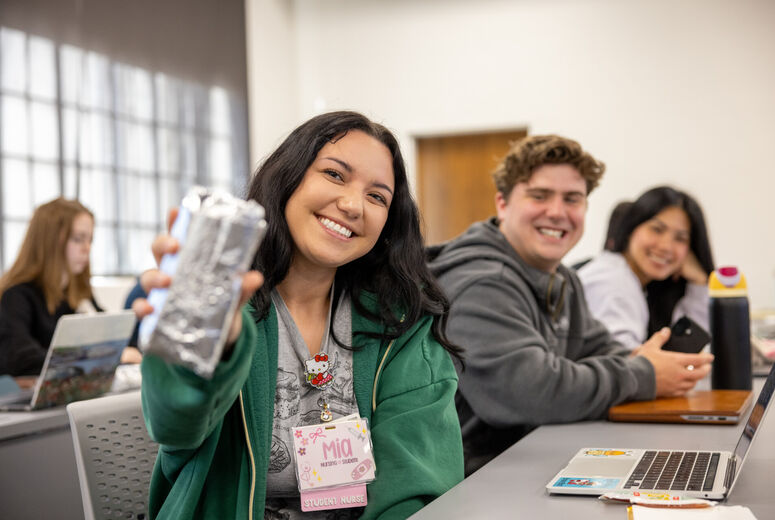 A smiling nursing student sits in a classroom and holds up a wrapped burrito toward the camera while classmates sit at desks in the background using laptops. The student wears a green hoodie. The classroom is bright and informal, with several students smiling and interacting during class.