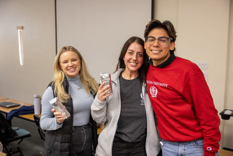 Three nursing students stand indoors in a classroom or meeting room, holding foil‑wrapped burritos, with desks and chairs visible in the background.