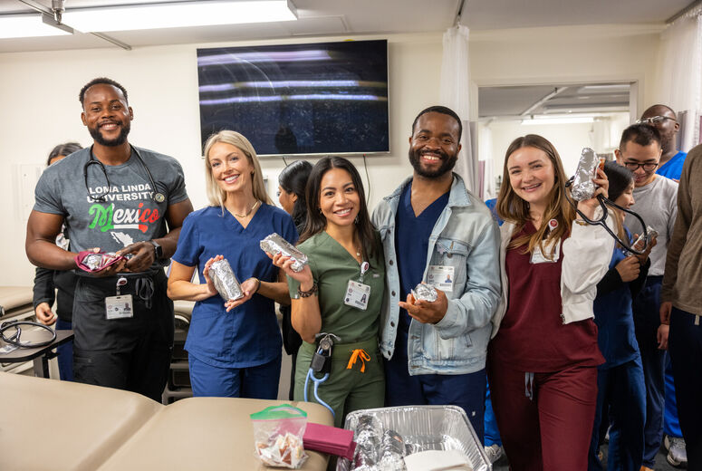 A group of nursing students stand together inside a clinical skills or healthcare training room, holding foil‑wrapped food items, with medical equipment, tables, and a wall‑mounted screen behind them.