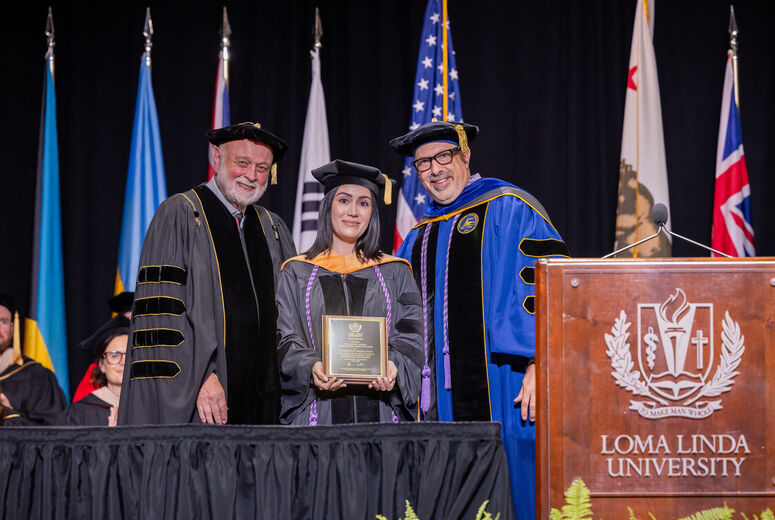 Female graduate stands between two administrators next to a podium holding a plaque in her hands. 