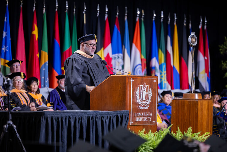 Commencement speaker stands at a podium addressing graduates as a panel of faculty and administrators sit behind him. 
