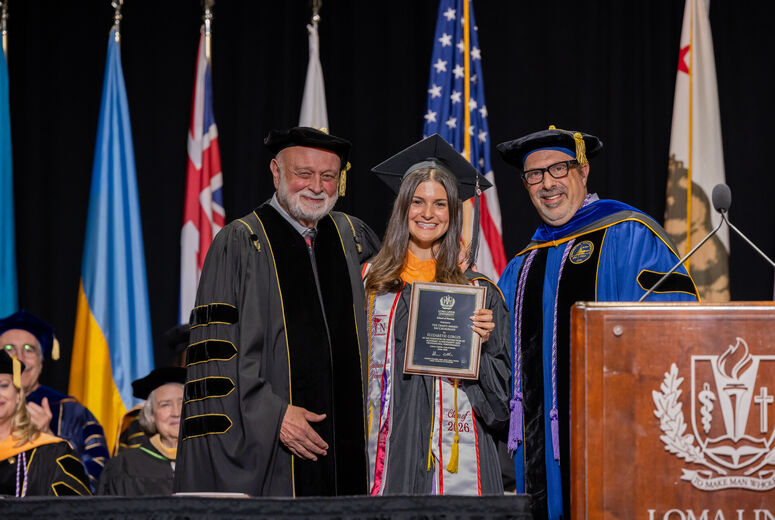 Female graduate stands between two administrators next to a podium holding a plaque in her hands. 