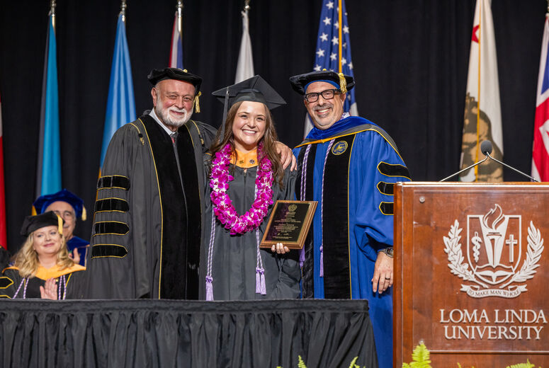 Female graduate stands between two administrators next to a podium holding a plaque in her hands. 